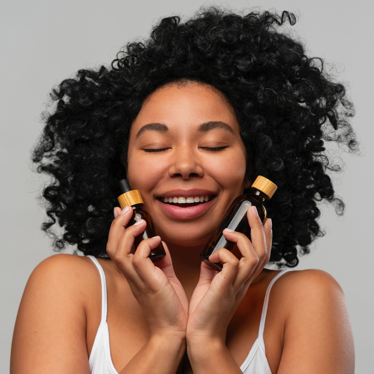 Joyful woman with natural curls holding two skincare bottles to her cheeks